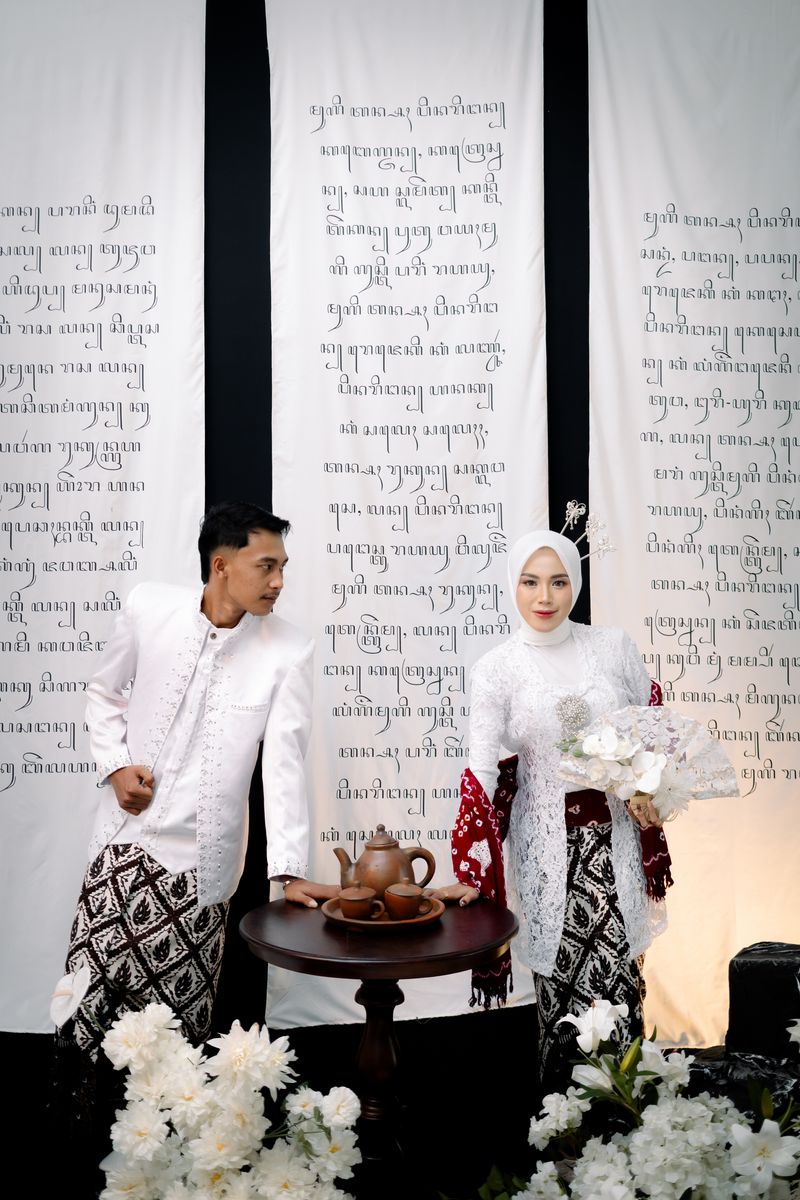 Pasangan berpose di samping meja teh dengan latar aksara Jawa