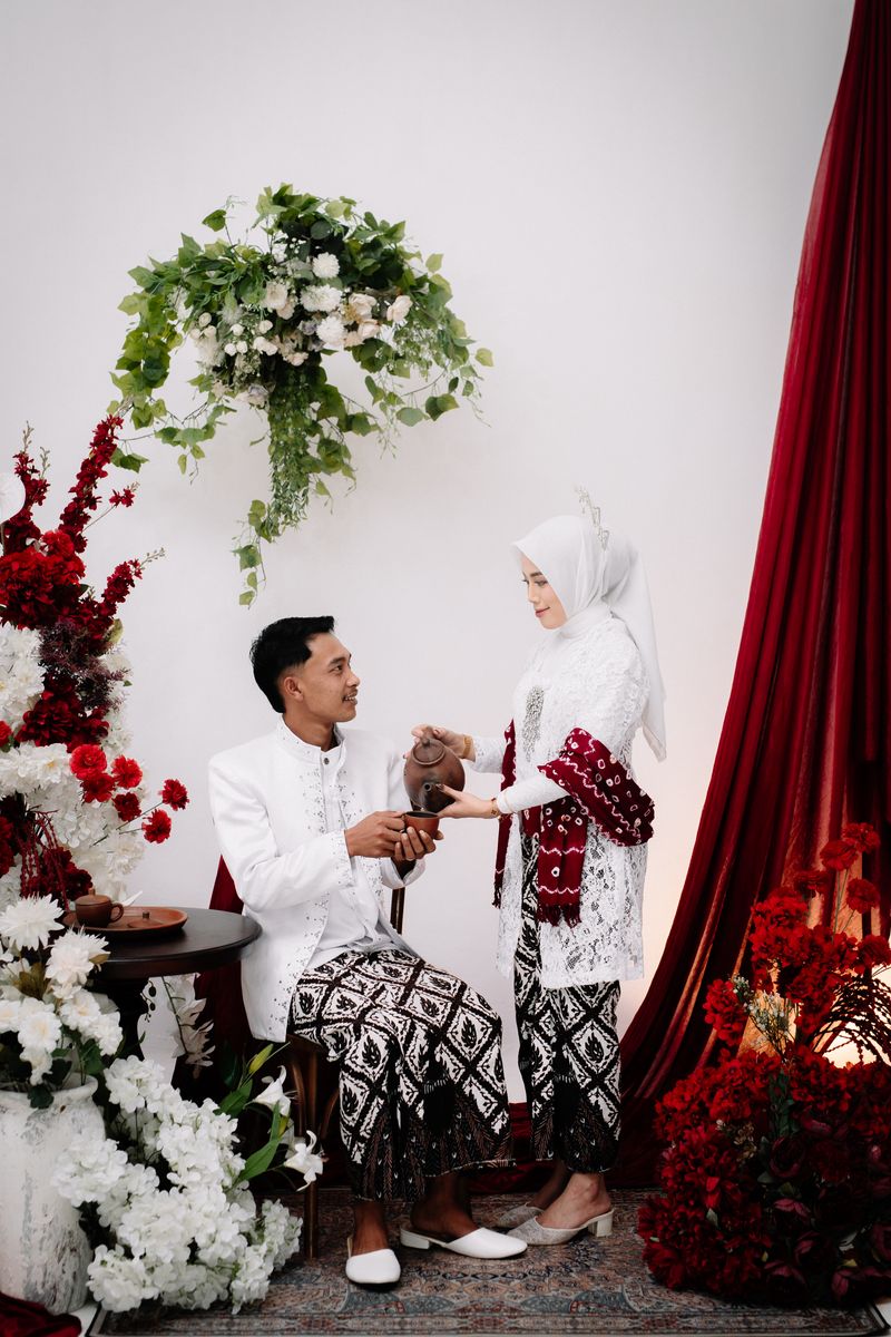 Safrina menuangkan teh untuk Rahardiyan di antara bunga dan tirai burgundy